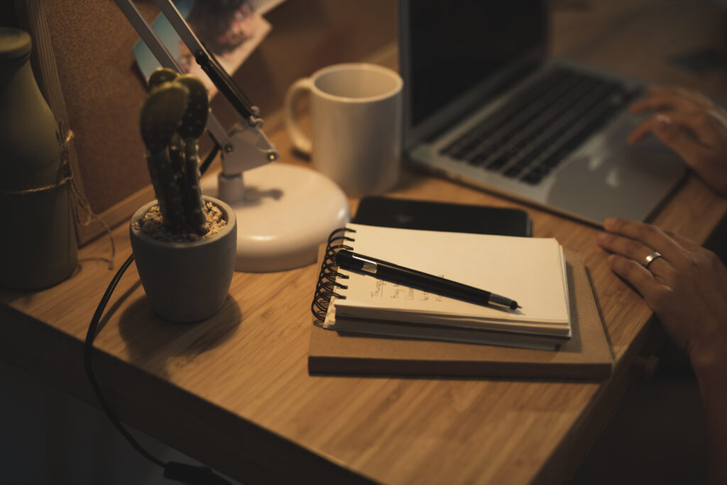 Image of a desk with a laptop besides some notebooks and notes, and someone probably working.